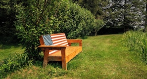 A wooden bench with an American flag cushion in a grassy park area. - Private Log Cabin Retreat with all Amenities in Greene, NY. (Greene)