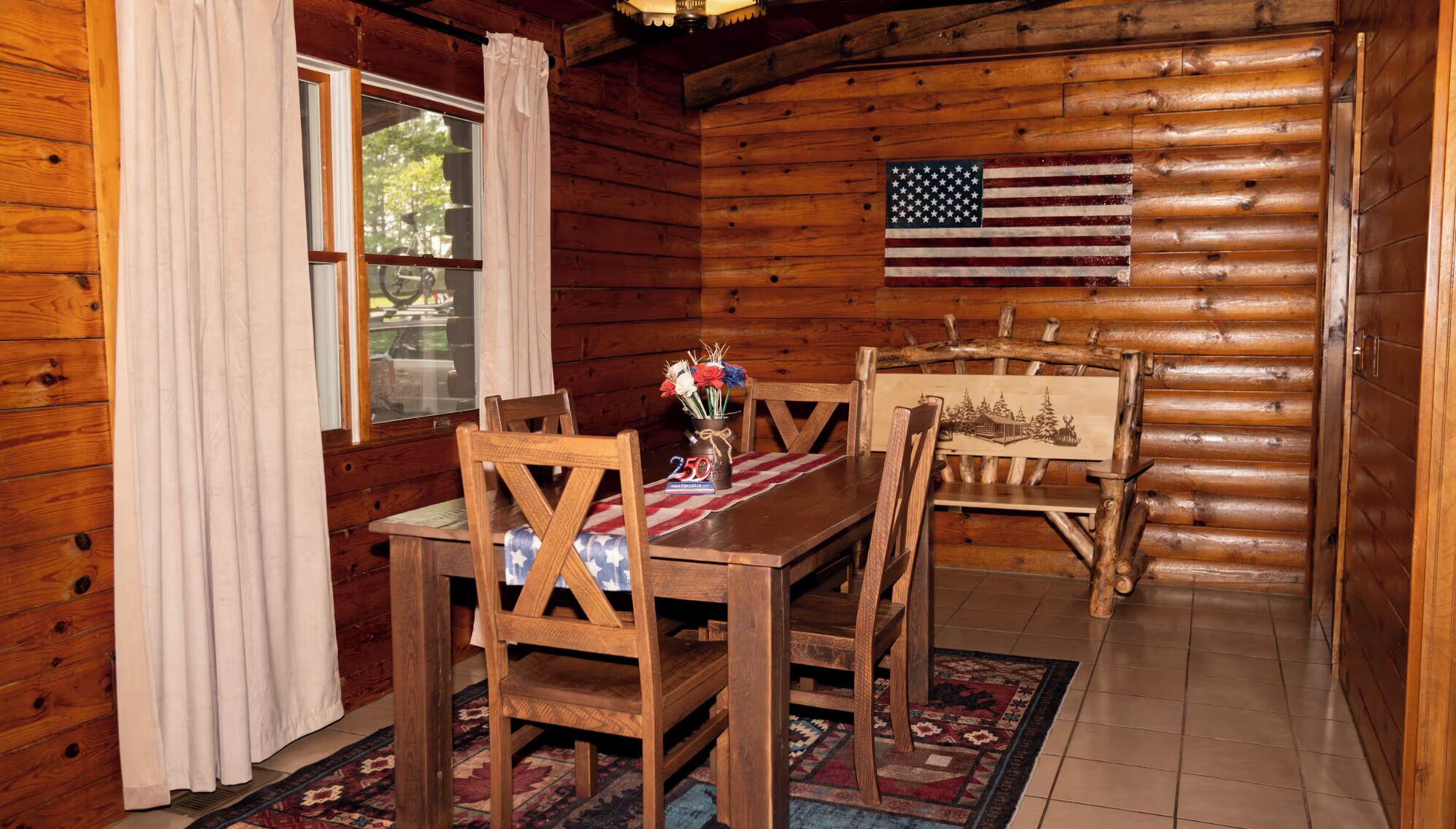 L’intérieur d’un chalet en bois, avec une table à manger, des chaises, un drapeau et une fenêtre avec des rideaux.