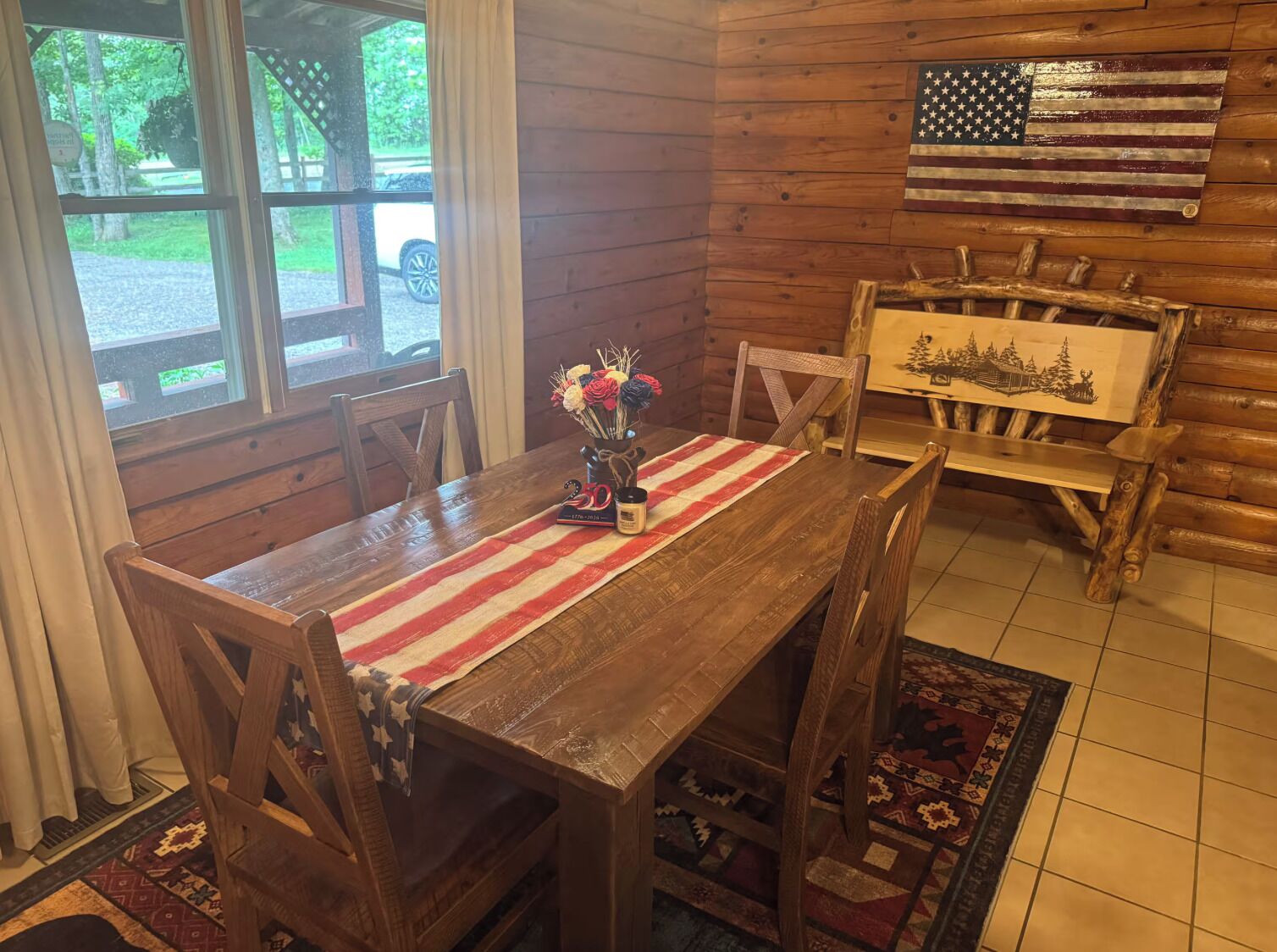 Un coin repas en bois avec une table dressée pour deux personnes, un drapeau accroché au mur et une vue sur l’extérieur.