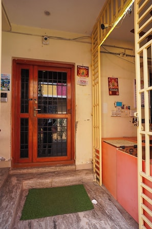 A wooden door with a metal grill, a small mat at the entrance, and a wall with various posters and electrical outlets. - Vel Ohm Homestay (Puducherry)