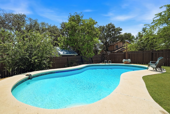 A swimming pool with a curved edge, surrounded by a wooden fence and trees. - Pool Haven | Hot Tub Fire Pit Game Room Sleeps 18 (San Antonio)