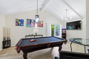 A pool table with billiard balls, a billiard cue rack, and a table tennis table in a room with a high ceiling and artwork on the walls. - Pool Haven | Hot Tub Fire Pit Game Room Sleeps 18 (San Antonio)