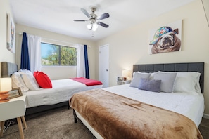 A hotel room with two beds, a ceiling fan, a window with curtains, a painting on the wall, and a nightstand with a lamp. - Pool Haven | Hot Tub Fire Pit Game Room Sleeps 18 (San Antonio)