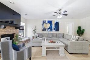A modern living room with a sectional sofa, a fireplace, a ceiling fan, and abstract wall art. - Pool Haven | Hot Tub Fire Pit Game Room Sleeps 18 (San Antonio)