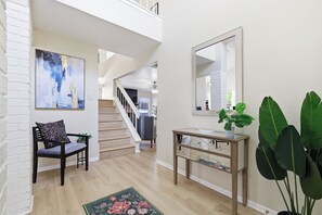 A spacious hallway with a staircase, a seating area with a chair and a rug, a console table with a mirror, and a large potted plant. - Pool Haven | Hot Tub Fire Pit Game Room Sleeps 18 (San Antonio)