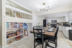 A modern kitchen with white cabinets, a dining area with a wooden table and chairs, and a bookshelf with various board games. - Pool Haven | Hot Tub Fire Pit Game Room Sleeps 18 (San Antonio)