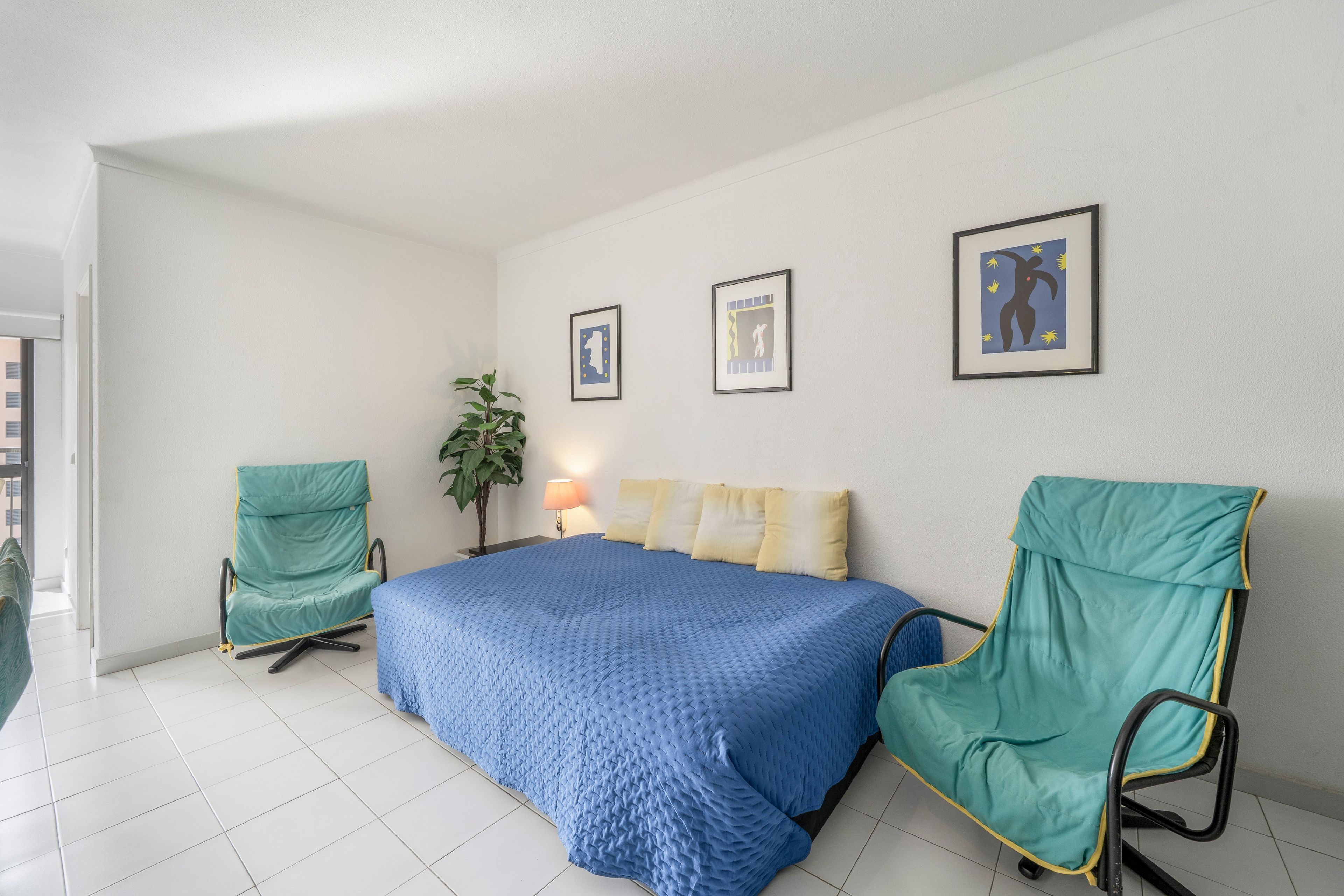 Un dormitorio con una cama, dos sillas, una lámpara y dos cuadros en la pared.