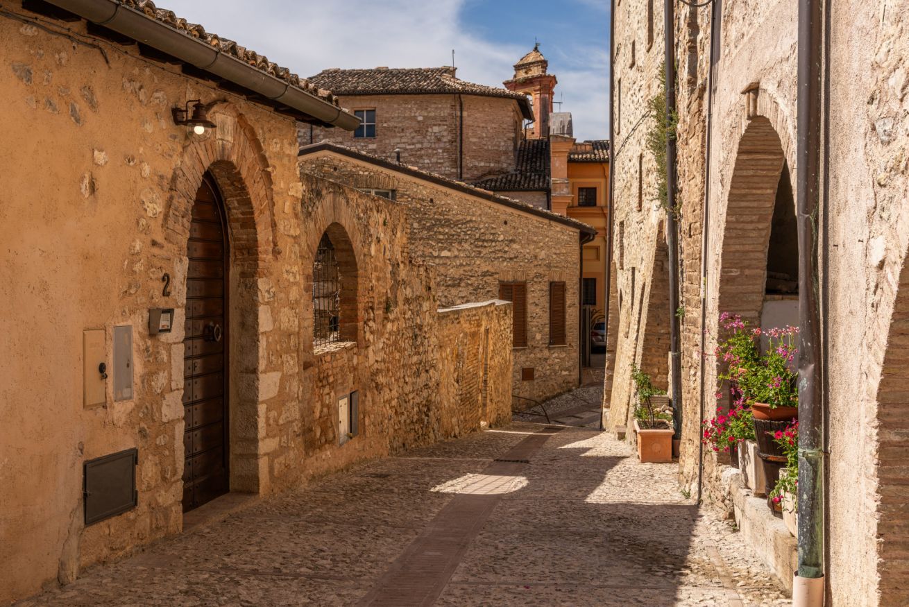 A narrow cobblestone street flanked by historic stone buildings with arched doorways and traditional tiled roofs.
