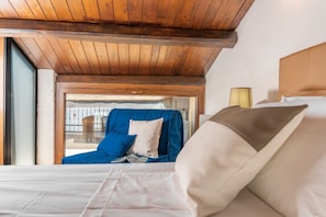 Habitación de hotel con techo de madera, cama con ropa de cama blanca, un sillón azul y vistas a un espacio exterior.