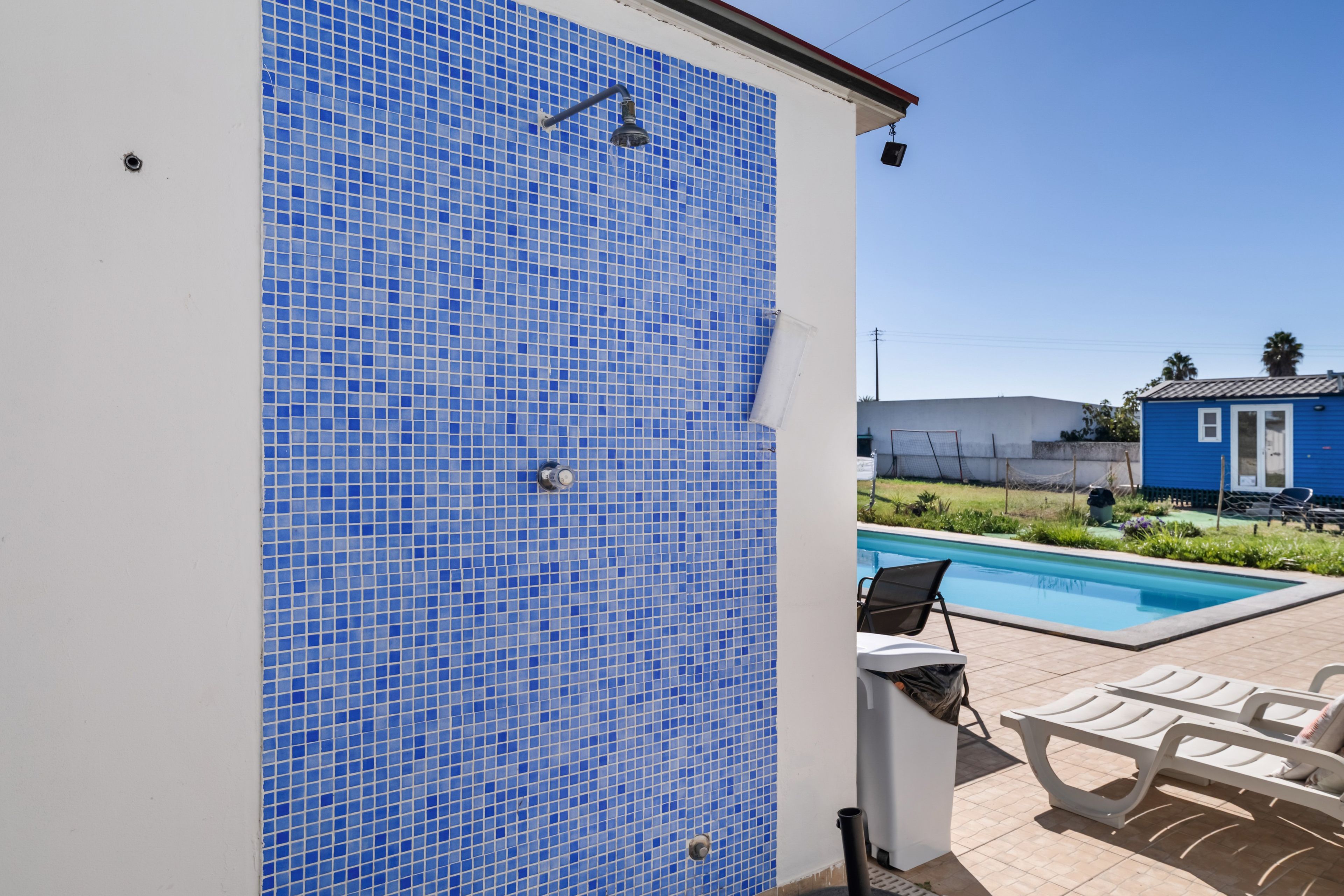 Ein Poolbereich mit einer blauen Fliesenwand, einer weißen Wand mit einer Dusche und einem wolkenlosen Himmel.