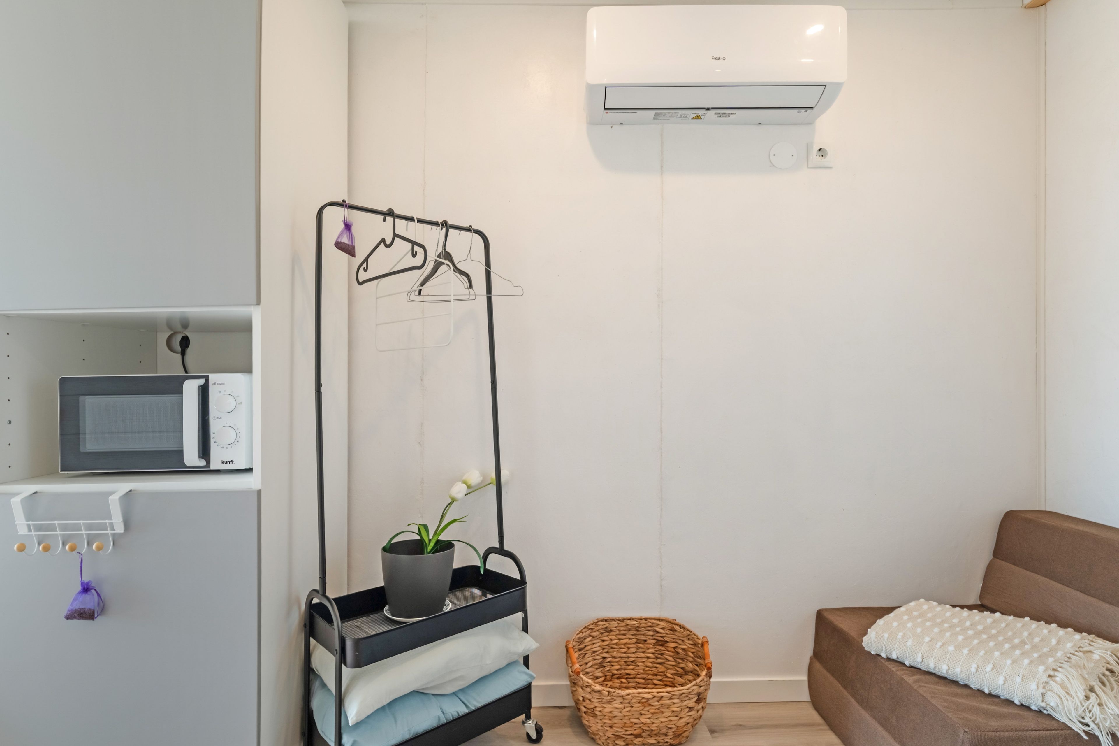 A modern, minimalist room with a built-in microwave, a wall-mounted air conditioner, a drying rack with clothes, a potted plant, a woven basket, and a cozy brown sofa with a white throw blanket.