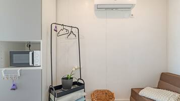 A modern, minimalist room with a built-in microwave, a wall-mounted air conditioner, a drying rack with clothes, a potted plant, a woven basket, and a cozy brown sofa with a white throw blanket.
