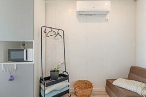 A modern, minimalist room with a built-in microwave, a wall-mounted air conditioner, a drying rack with clothes, a potted plant, a woven basket, and a cozy brown sofa with a white throw blanket.