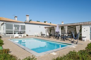 A swimming pool with lounge chairs and umbrellas, a building with large windows, and a tiled roof.