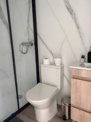 A modern bathroom with a toilet, a glass shower enclosure, a sink, and a small trash bin. - Tiny House with Shared Pool, Wi-Fi, and Air Conditioning (Palmela)