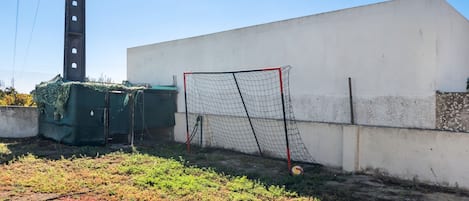 Un but de football situé dans un espace délimité par une clôture, avec un mur blanc et une structure recouverte d’une bâche verte.