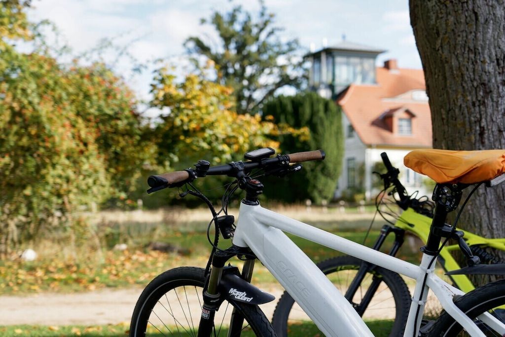 En vit cykel med en monterad anordning, lutad mot ett träd, med ett hus och grönska i bakgrunden.