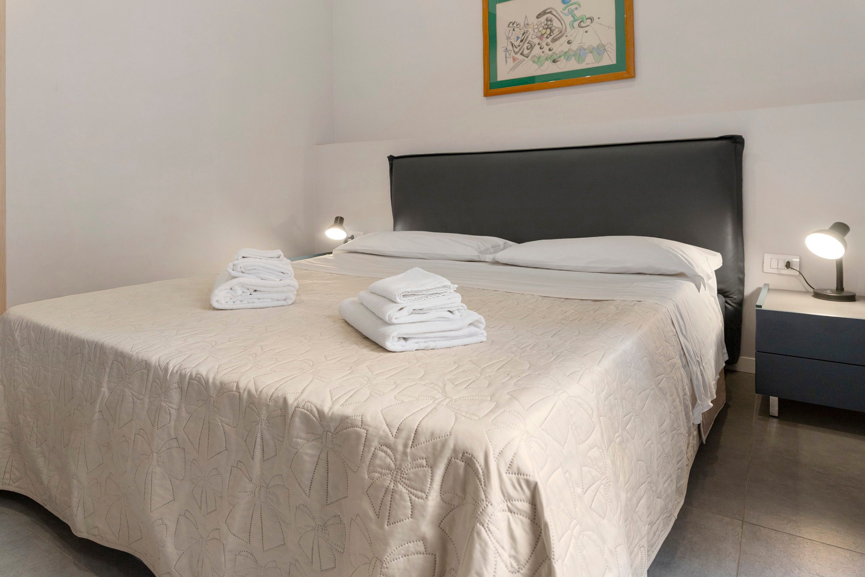 A neatly made bed with a quilted bedspread, two bedside lamps, and a framed picture on the wall.