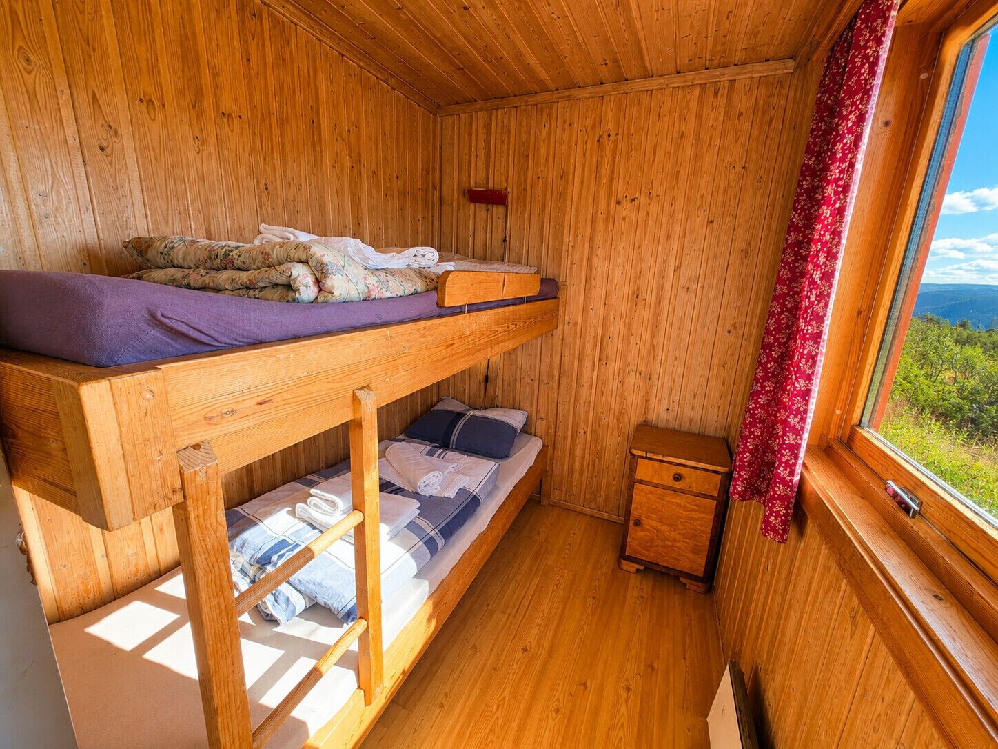 Eine kleine Holzhütte mit einem Etagenbett, einem kleinen Holzschrank und einem Fenster mit Blick auf Grünfläche.