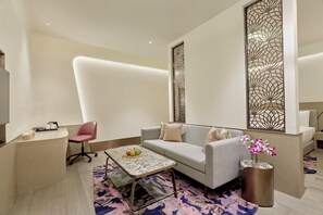 A modern living room with a grey sofa, a pink office chair, a coffee table, and a decorative wall panel. - Swirl Transit Hotel by Adani (Panvel)
