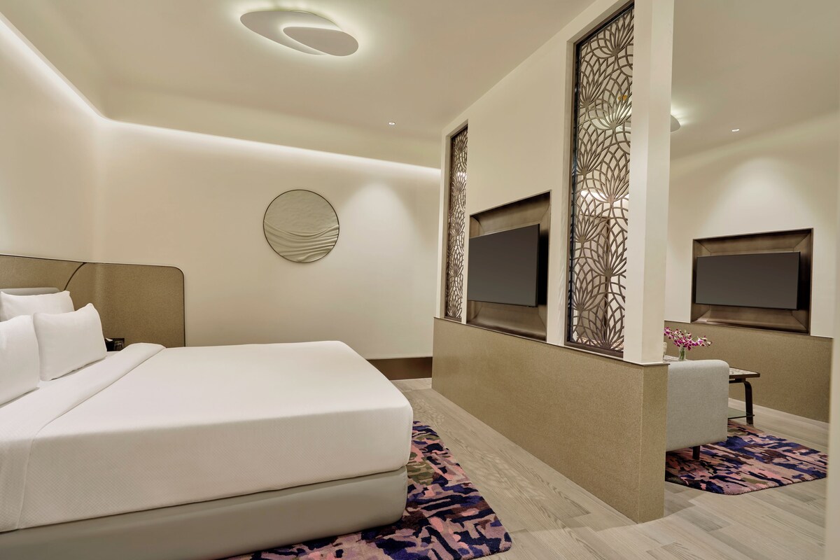A modern hotel room with a large bed, a flat-screen TV, a decorative wall panel, and a small table with a vase of flowers.