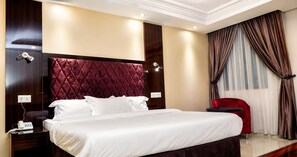 A hotel room with a large bed, a red upholstered chair, a bedside table with a phone, and a window with curtains. - Starview Palace Abuja (Gwarinpa)