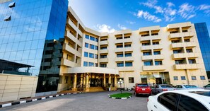 A multi-story hotel with a central entrance, a parking area with several cars, and a modern glass building adjacent to it. - Starview Palace Abuja (Gwarinpa)