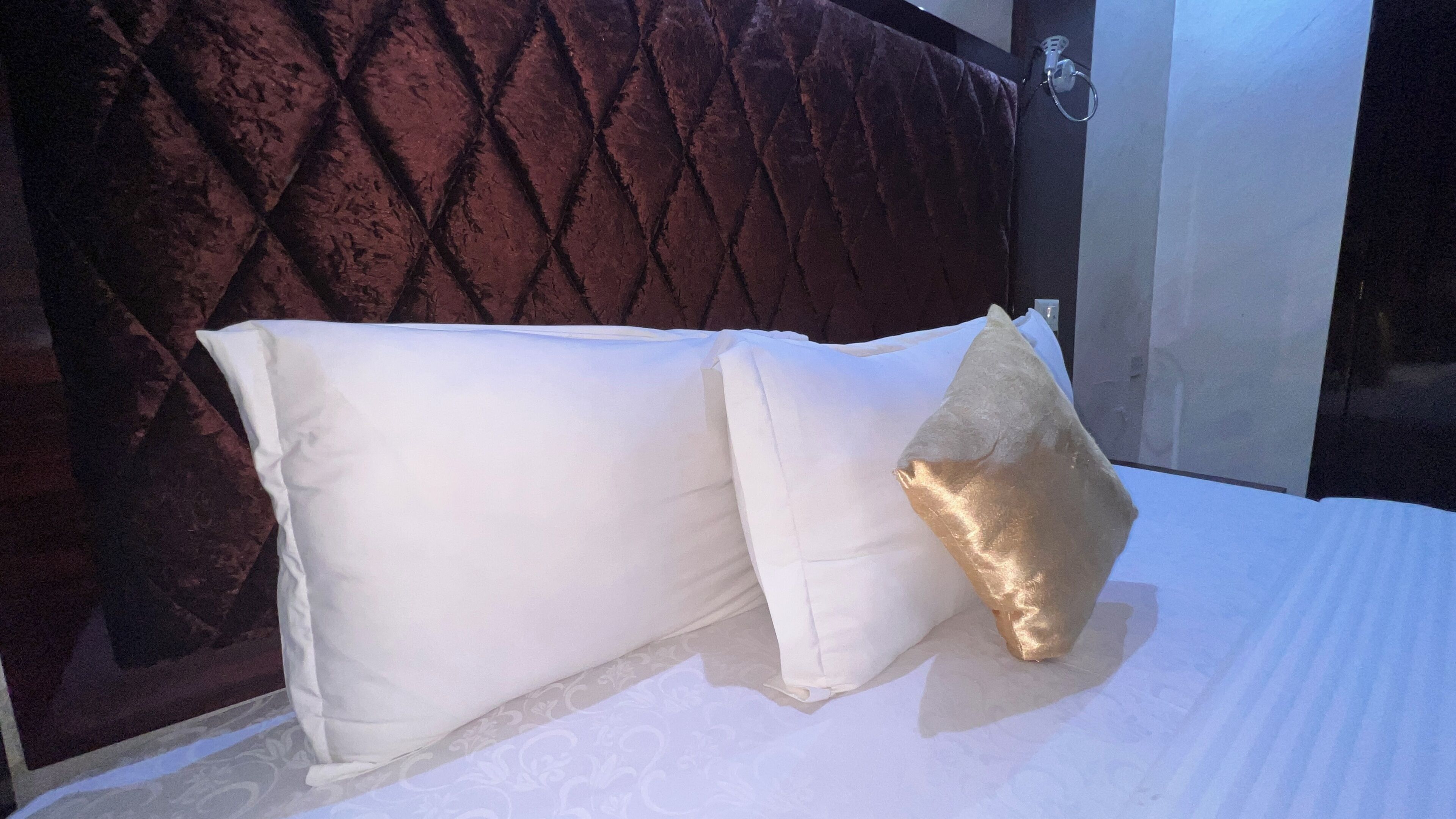Un lit avec des oreillers blancs et dorés, une tête de lit sombre et une porte de salle de bain visible.
