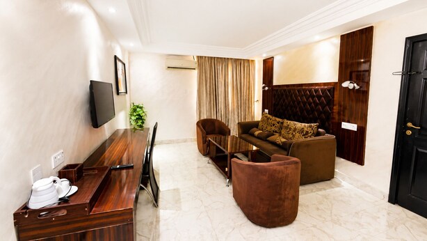 Quarto de hotel com mesa de jantar de madeira, um sofá com almofadas estampadas, poltrona marrom e porta preta.
