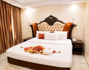 A hotel room with a large bed, a headboard with intricate carvings, a bedside table with a lamp, and a curtain on the window. - Starview Palace Abuja (Gwarinpa)