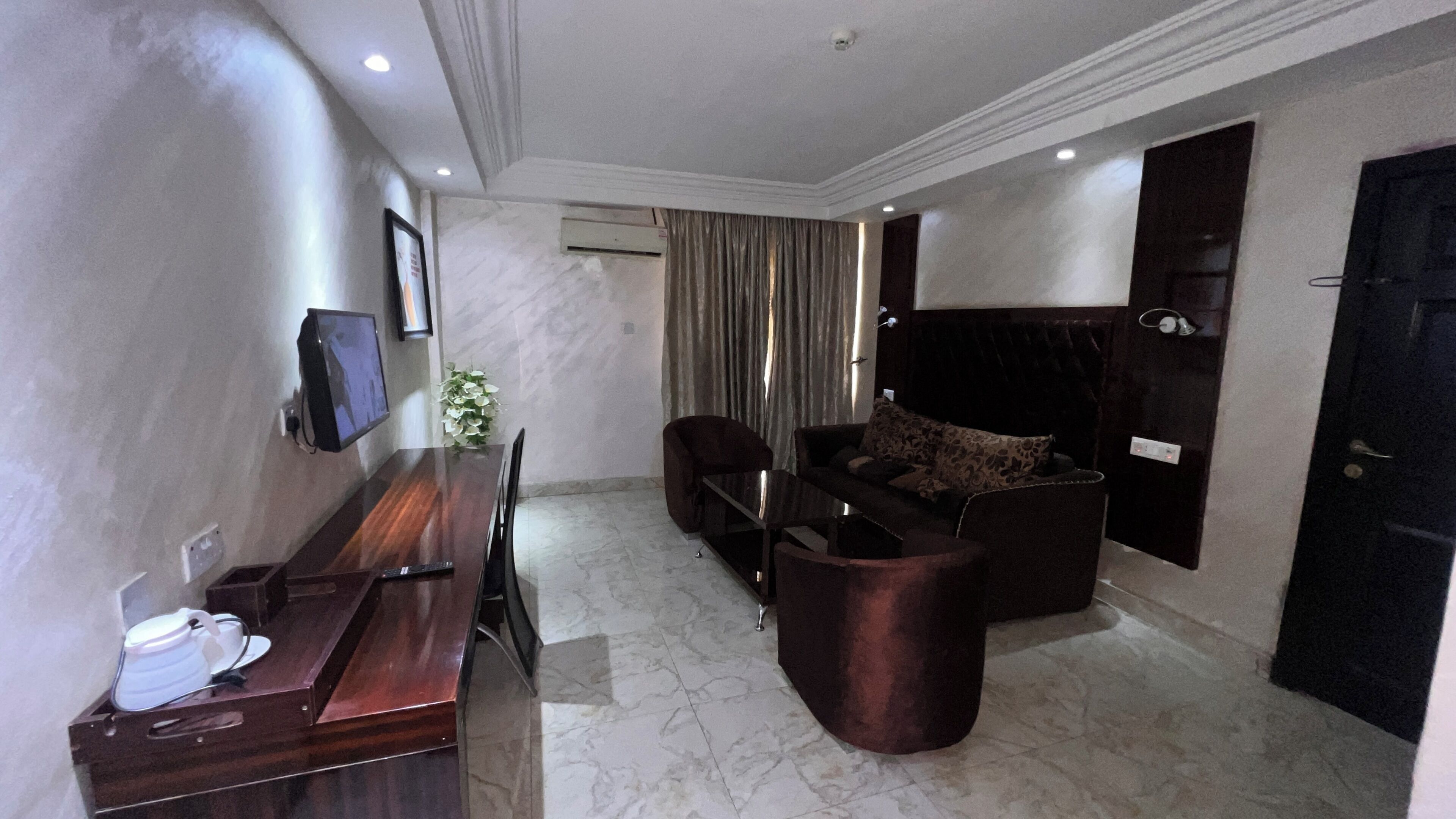 Quarto de hotel com uma mesa de jantar de madeira, um sofá, uma TV fixada na parede e uma porta com puxador.