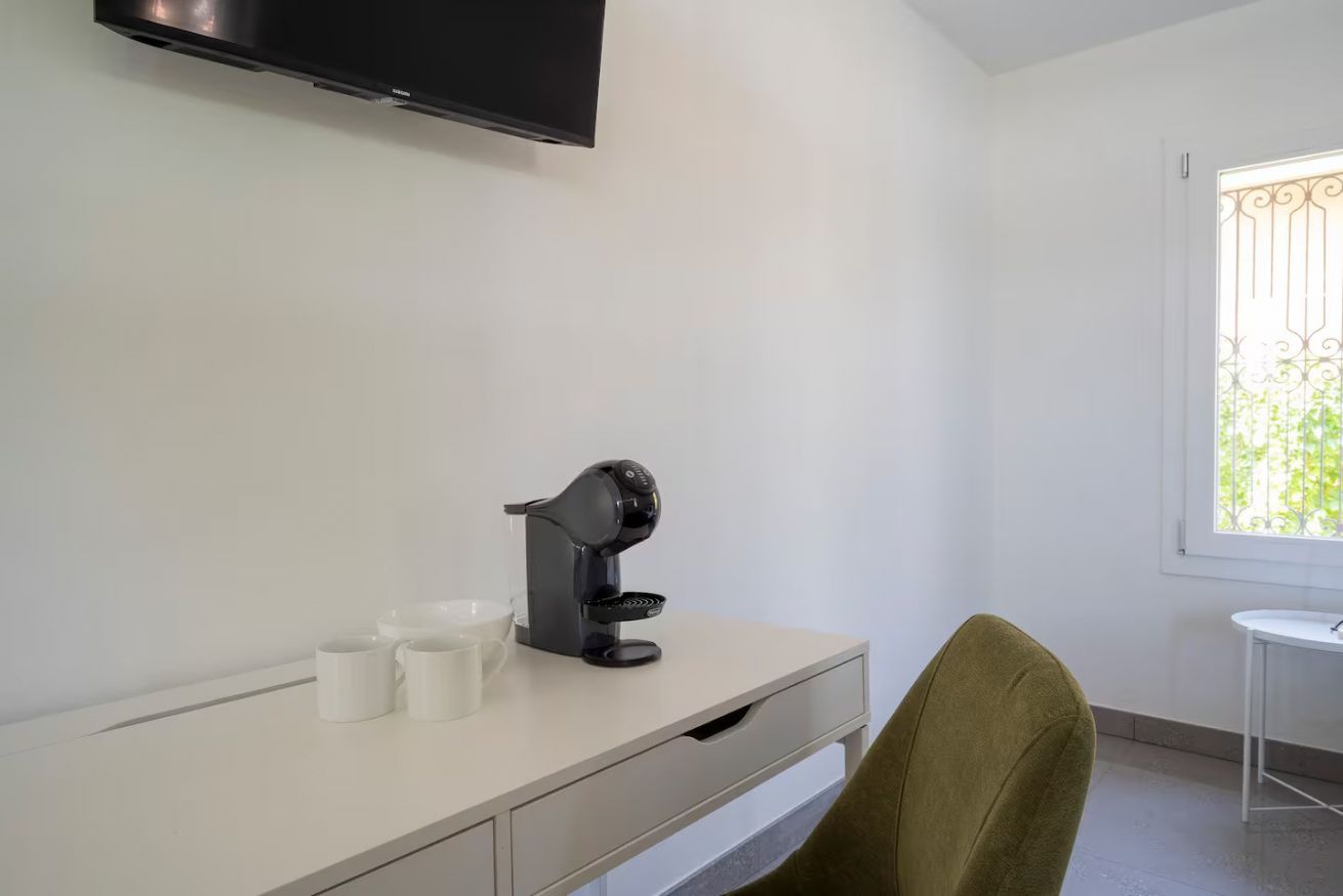 A room with a white desk, a black coffee machine, two white cups, a green chair, and a television mounted on the wall.