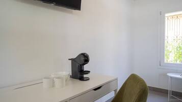 A room with a white desk, a black coffee machine, two white cups, a green chair, and a television mounted on the wall.
