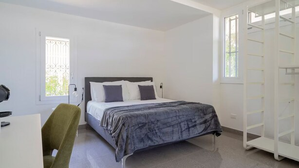 Ein Schlafzimmer mit einem Bett, einem Schreibtisch, einem Stuhl, einem Kleiderschrank und einem Fenster mit einem Metallgitter.