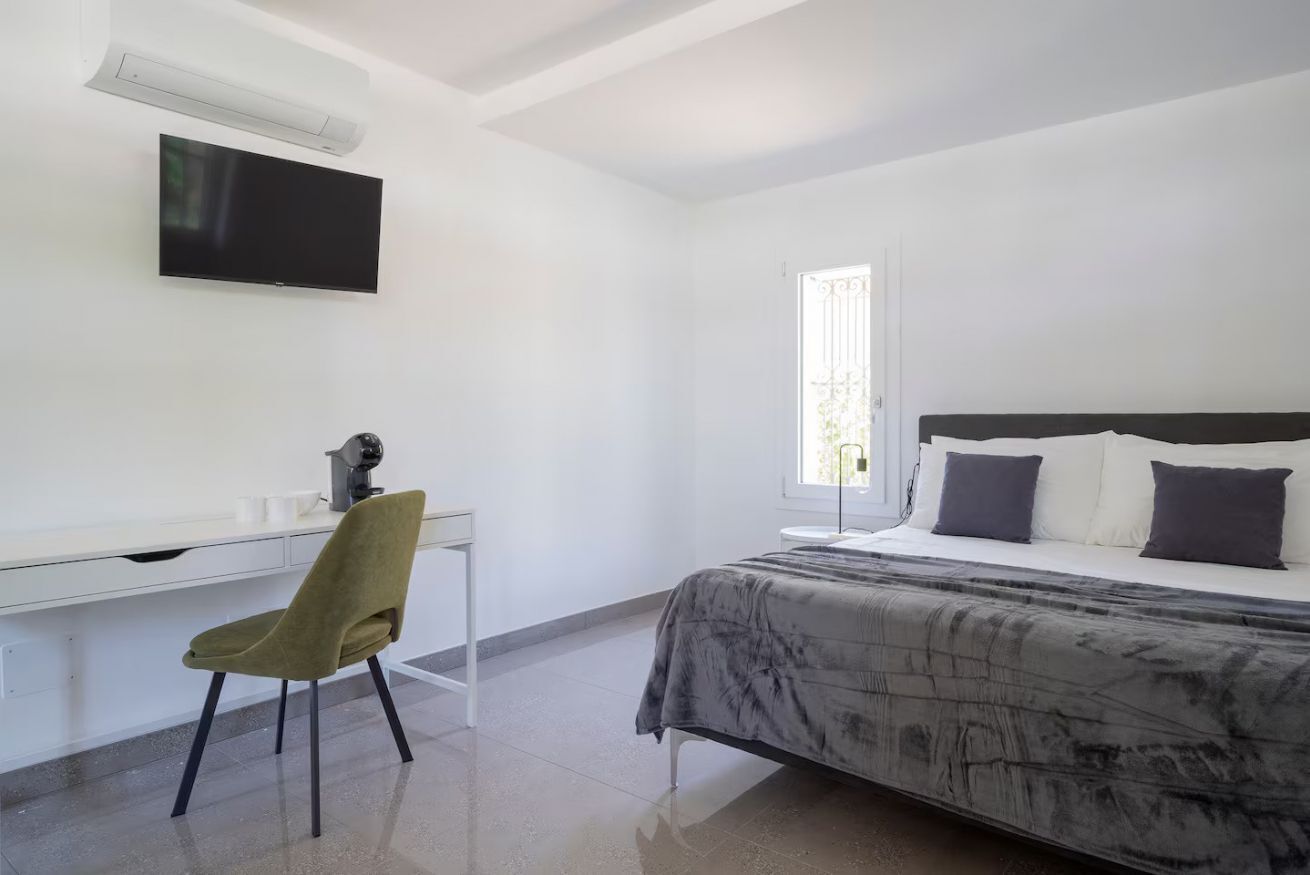 Quarto com cama, escrivaninha, cadeira, televisão e janela.