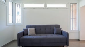 Ein modernes Wohnzimmer mit einem grauen Sofa, zwei Fenstern und einer schlichten weißen Wand.