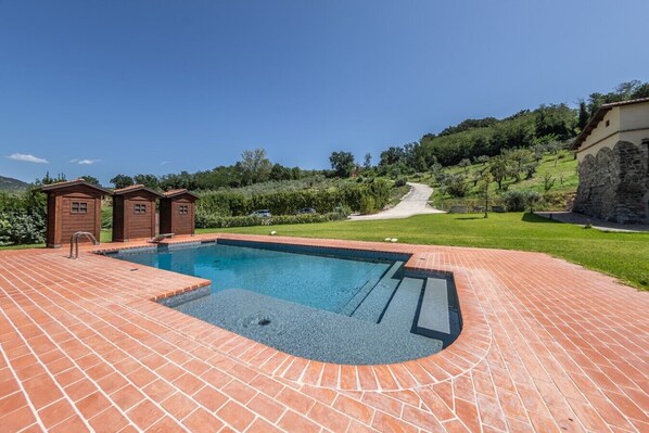A swimming pool with a brick deck, two wooden cabins, and a stone building in a landscaped garden.