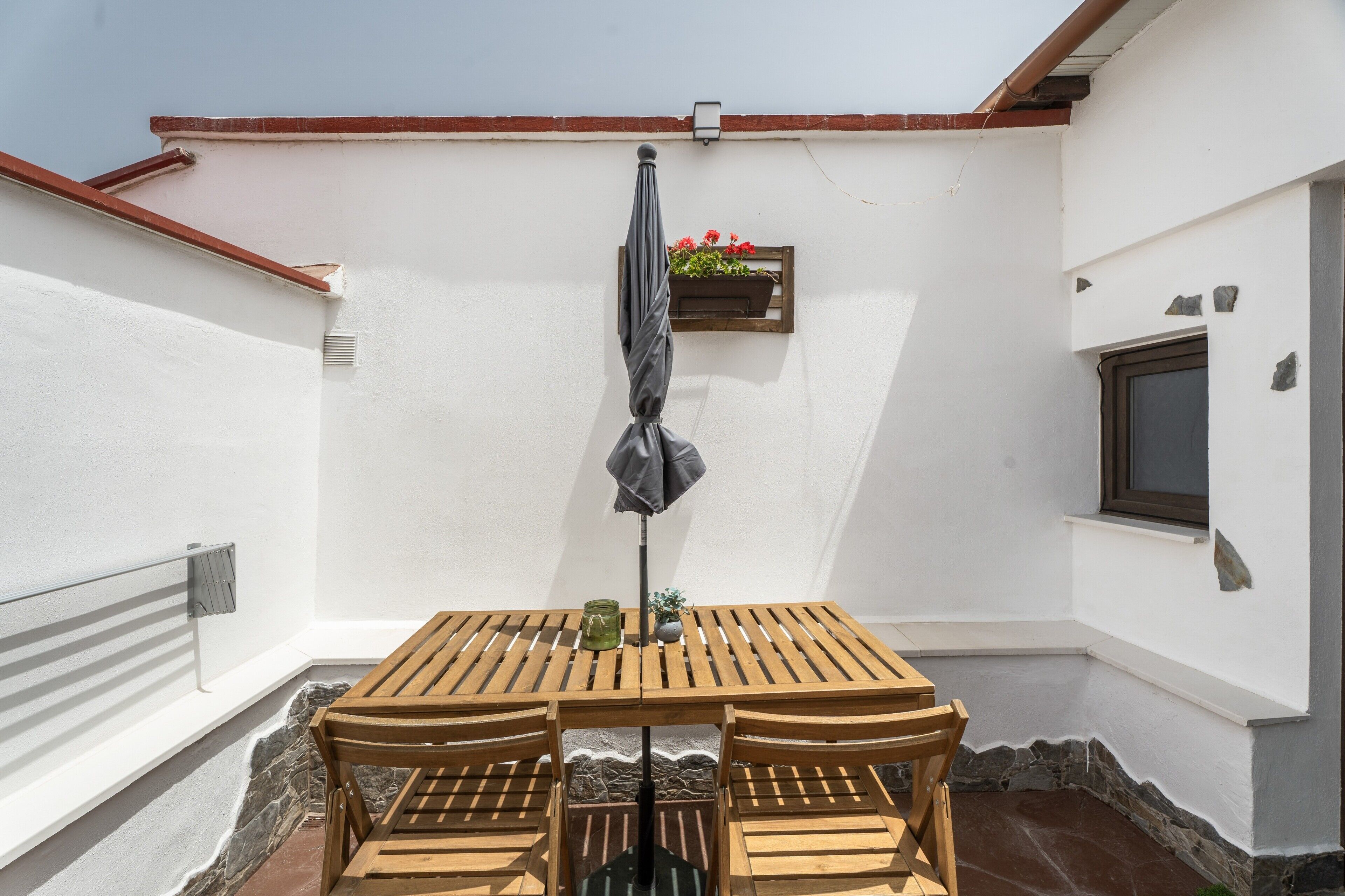 Ein Holztisch mit Stühlen steht auf einer Terrasse mit weißer Wand und Fenster.