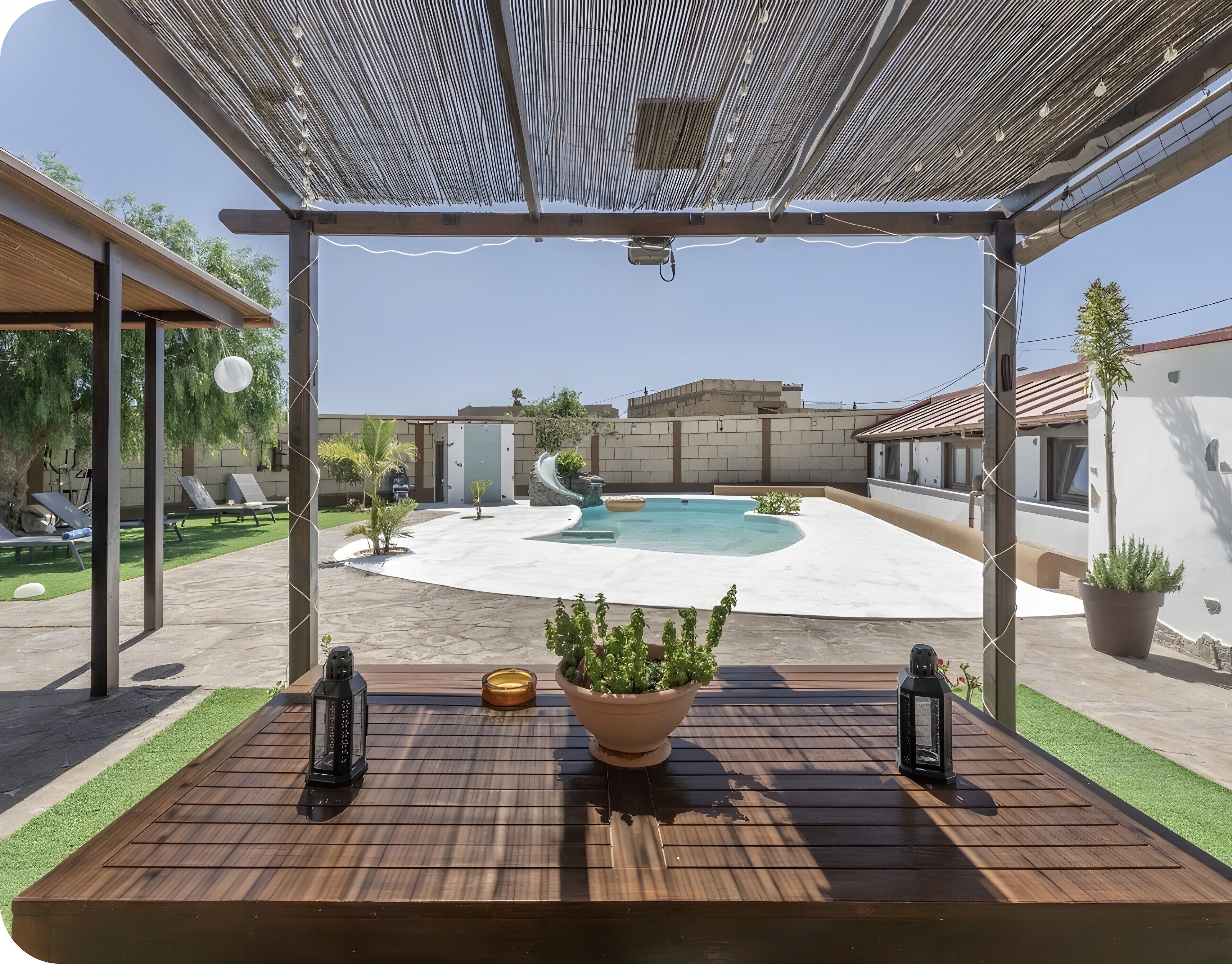 Eine Holzterrasse mit einem Blumentopf und zwei Laternen, mit Blick auf einen Swimmingpool und ein Gebäude.
