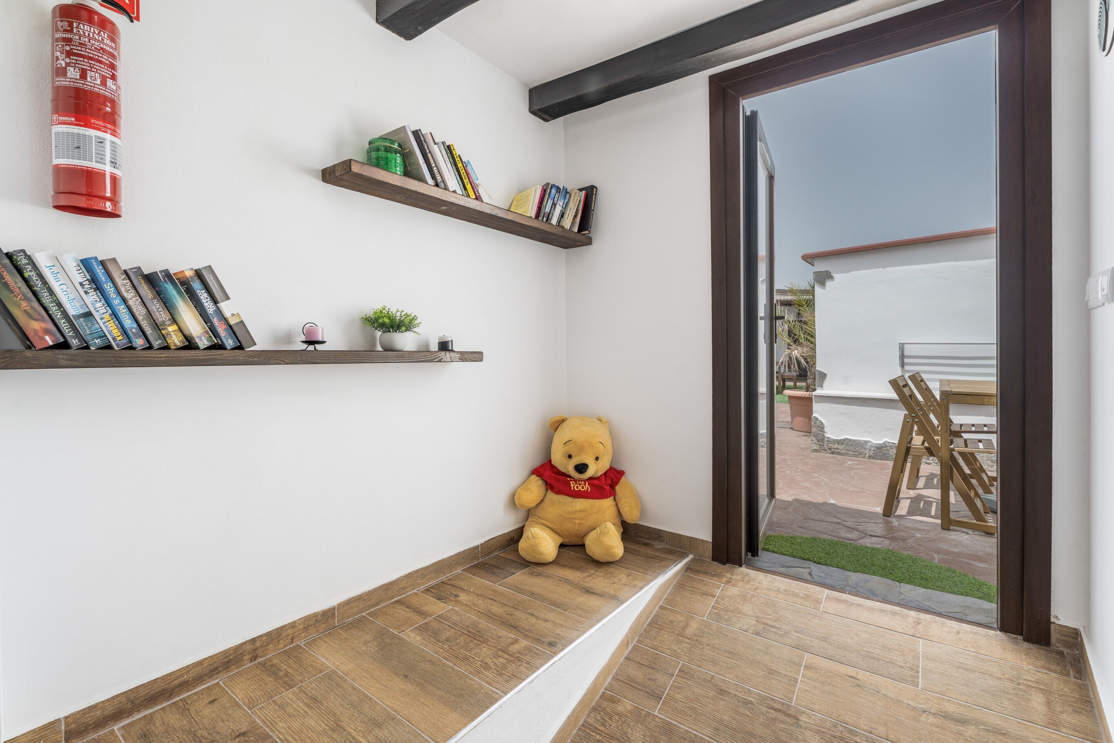 Quarto com piso de madeira, um urso de pelúcia e duas prateleiras com livros e uma pequena planta.