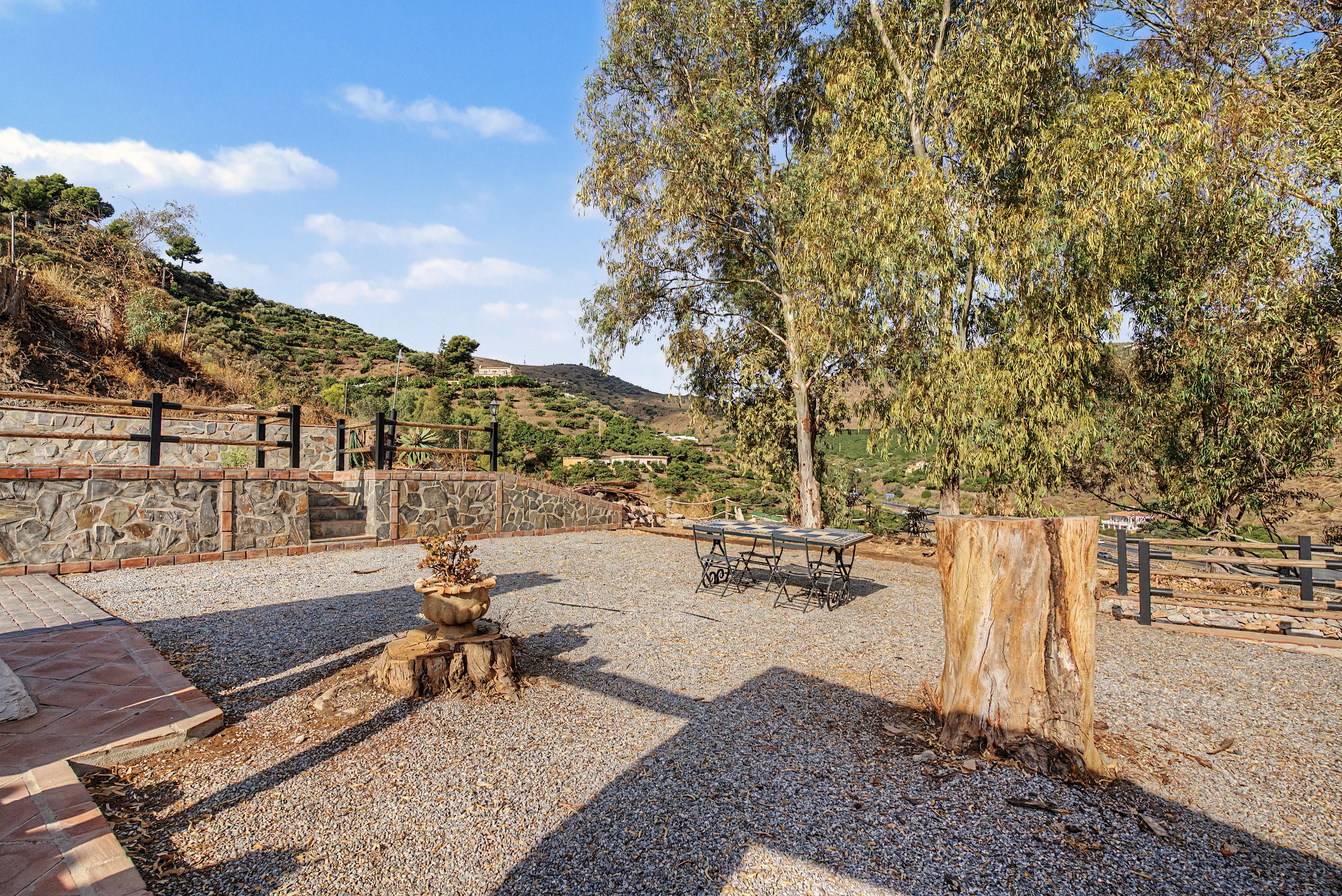 Un patio de grava con pared de piedra, una mesa de madera y bancos.