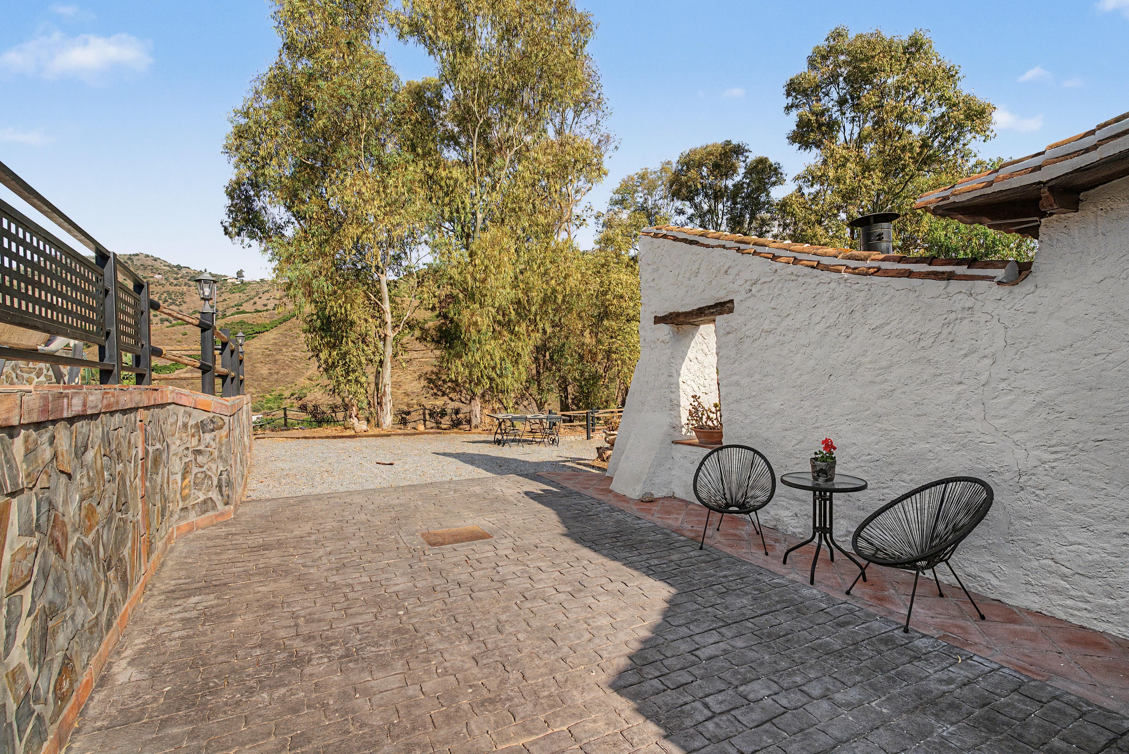 Un patio con muro de piedra, cerca metálica y edificio blanco con tejado de tejas. Hay dos sillas, una mesa y una planta en maceta en la pared.
