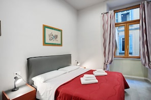 A hotel room with a bed, a nightstand, a desk lamp, a framed picture on the wall, and a window with curtains. - Double Room with External Bathroom, Wi-Fi and Air Conditioning (Napoli)