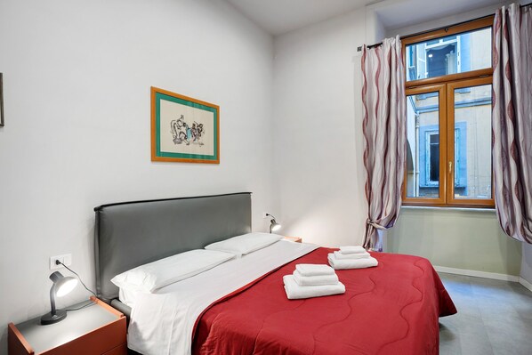 Une chambre d’hôtel avec un lit, une table de chevet, une lampe de bureau, un tableau encadré au mur et une fenêtre avec des rideaux.