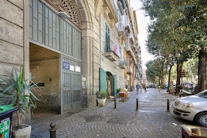 Une rue pavée bordée d’immeubles, avec une voiture garée et quelques piétons.