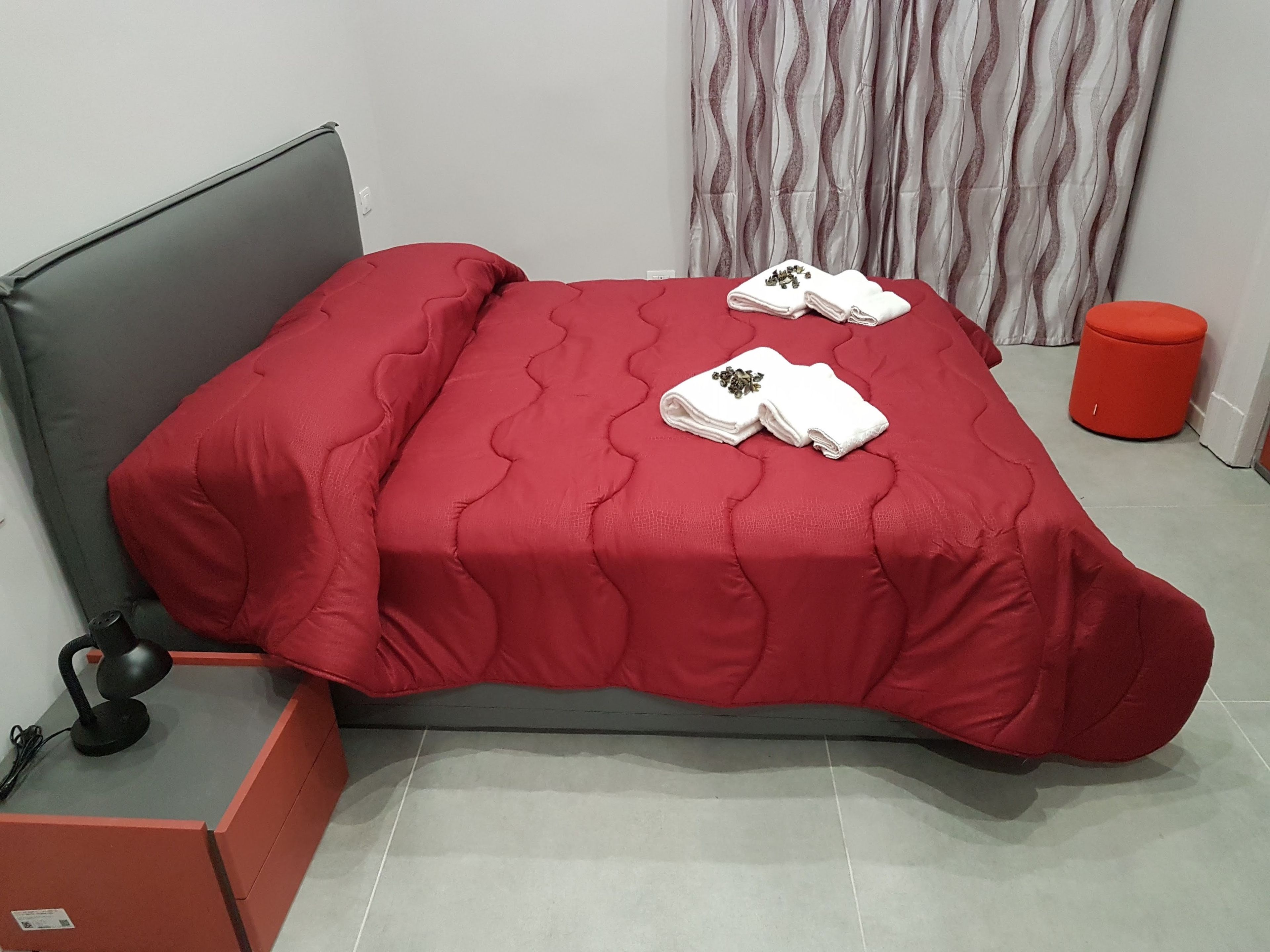 A bed with a red bedspread, two white towels with a floral design, a red cylindrical object, and a grey headboard.