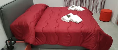 A bed with a red bedspread, two white towels with a floral design, a red cylindrical object, and a grey headboard.