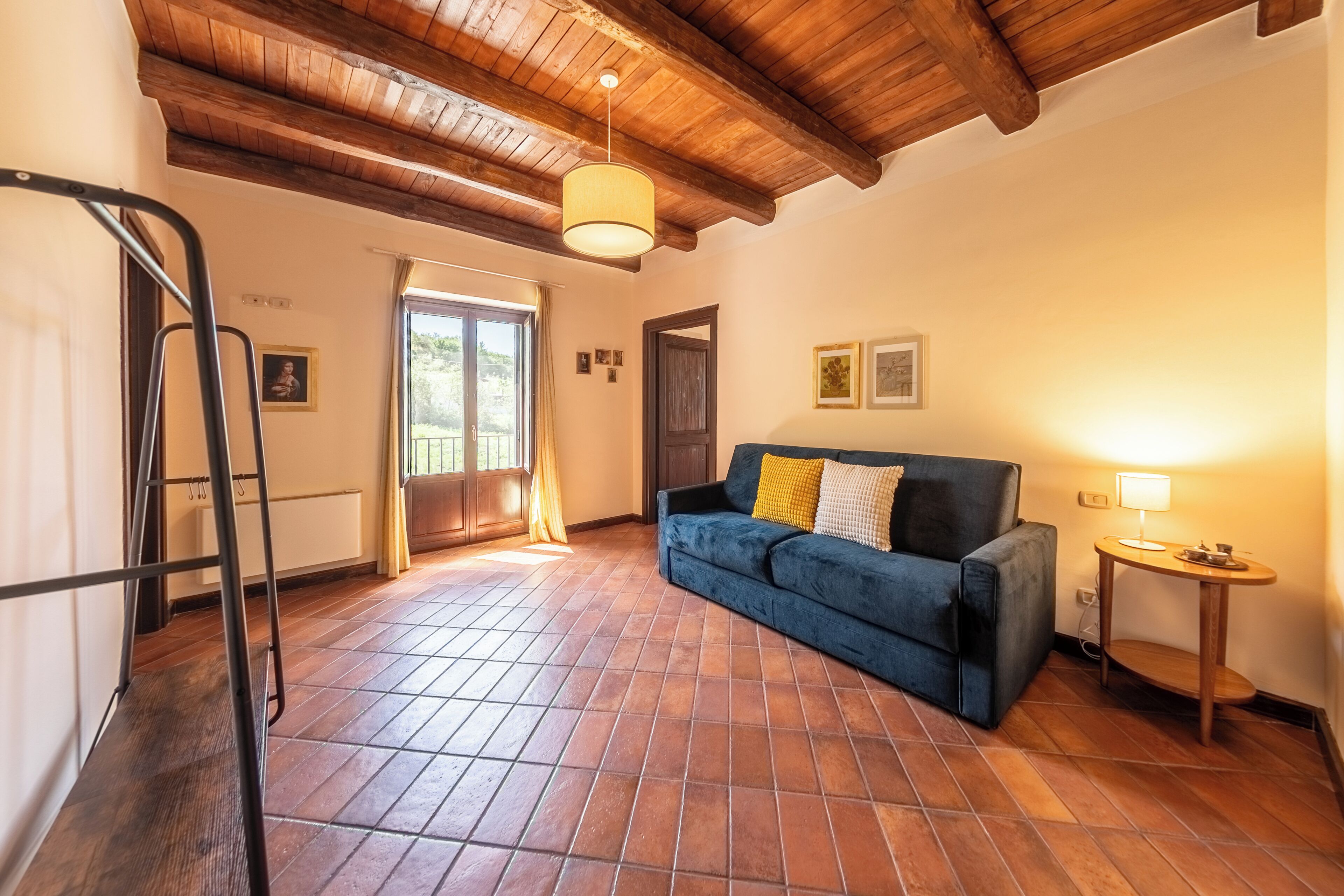 A room with a blue sofa, a small table with a lamp, a wooden ceiling, and a tiled floor.
