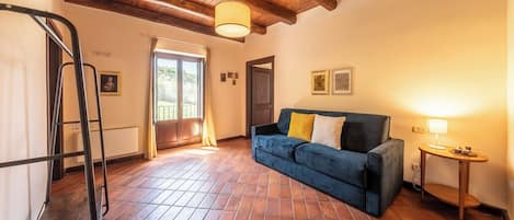 A room with a blue sofa, a small table with a lamp, a wooden ceiling, and a tiled floor.