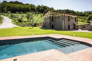 A pool area with a stone building, a paved path, and a grassy landscape with trees.